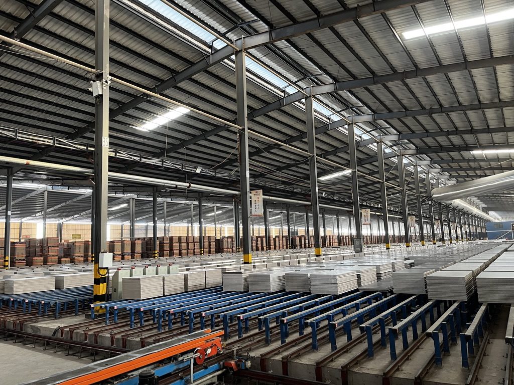 Finished tiles stacked on blue roller lines in the packing hall, warehouse cartons behind